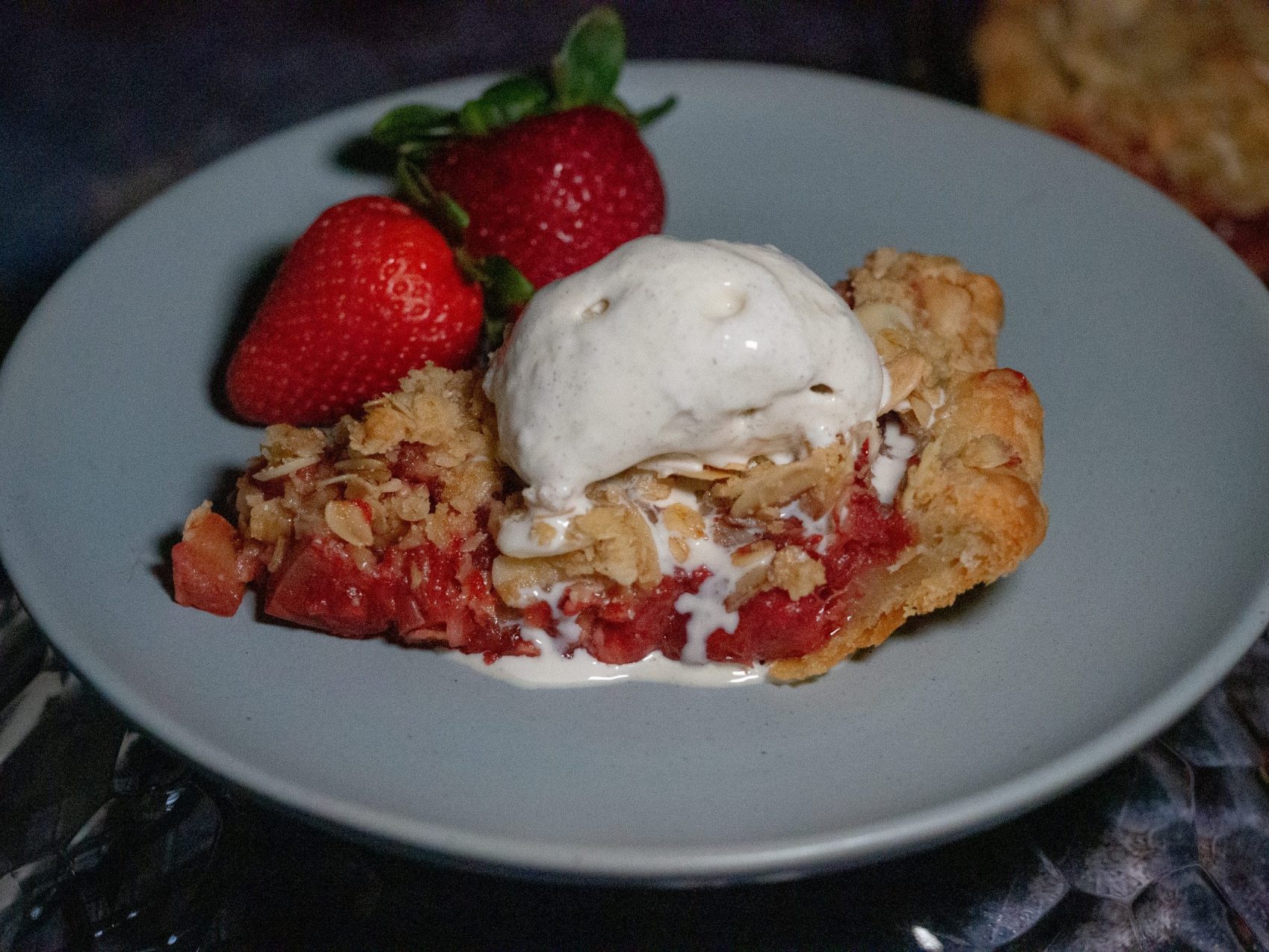 A slice of Strawberry-rhubarb pie crisp with vanilla ice cream and 2 fresh strawberries on a light blue plate