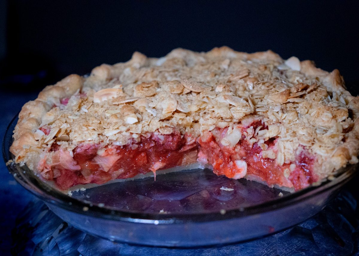 strawberry-rhubarb pie crisp with almond-oat streusel toppings on a dark grey patterned working space adorn with fresh strawberries.